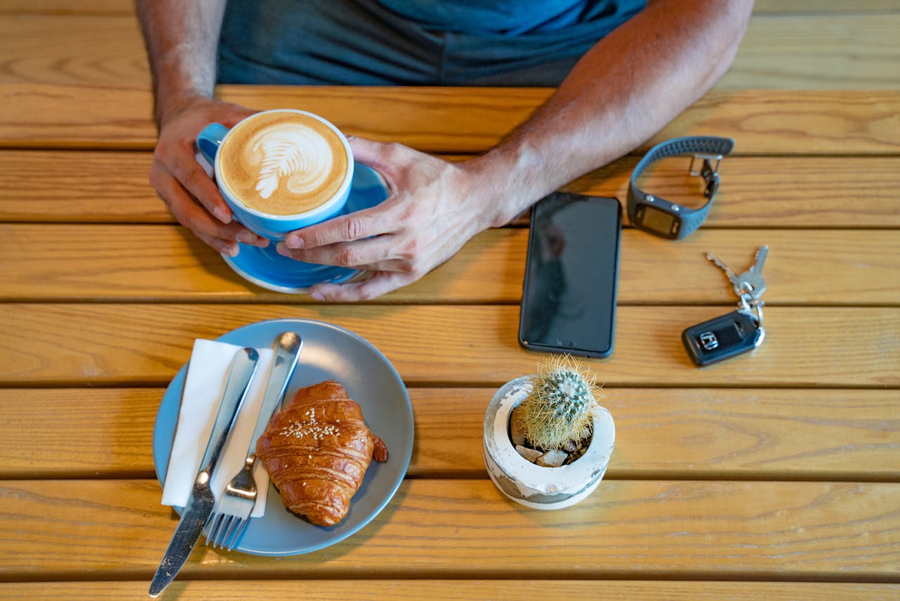 A cozy coffee setup with a croissant, latte, smartphone, wristwatch, and cactus.