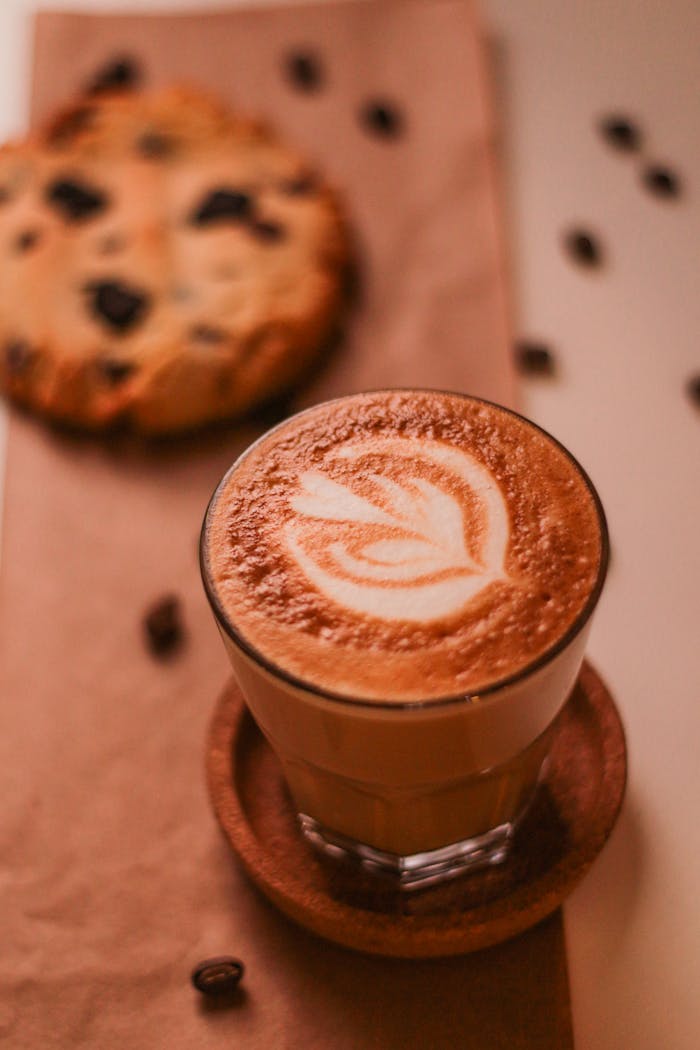 A delicious latte with art foam beside a chocolate chip cookie on a wooden table, perfect for coffee lovers.